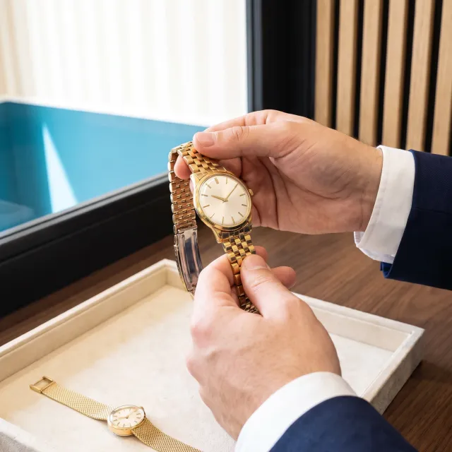 L’esperto esamina un orologio da uomo in oro con bracciale a maglia; sul vassoio, un orologio da donna in oro